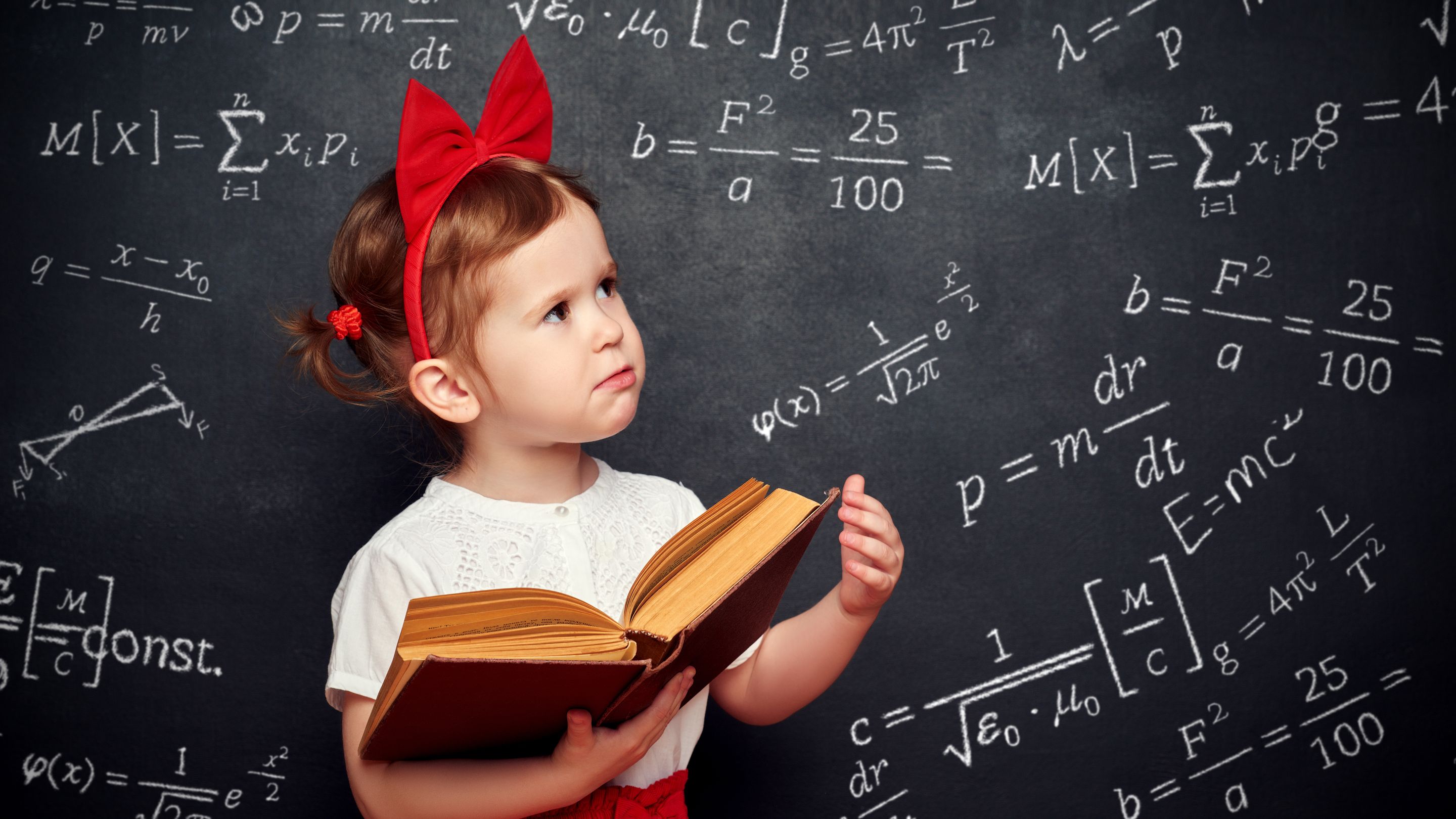 A young girl holding a book while in deep thought with a white board covered with math equations behind her.